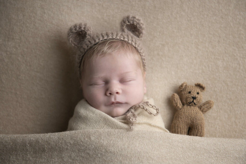 Newborn baby wrapped in a soft blanket wearing a teddy bear bonnet with a matching teddy